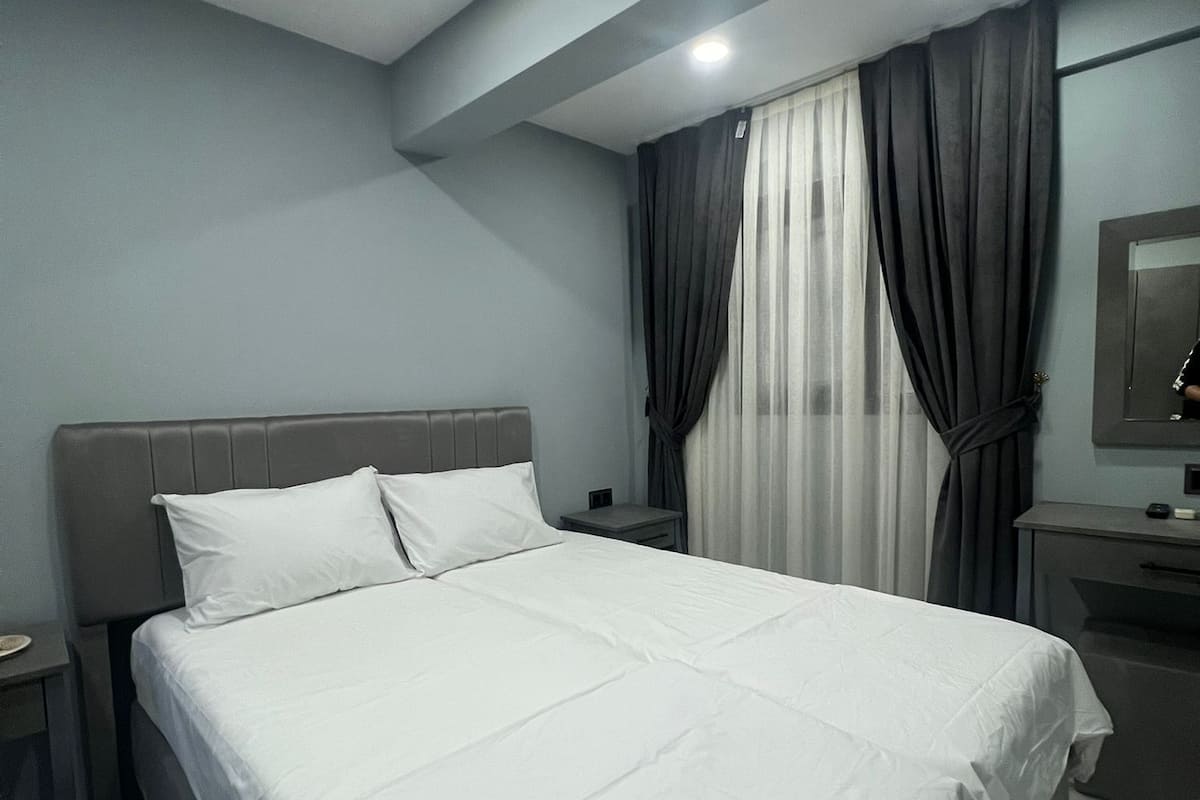 A modern hotel room with a large bed, a bedside table, a nightstand, a desk with a chair, and a mirror on the wall.