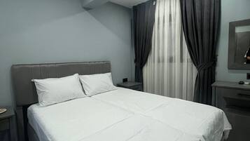A modern hotel room with a large bed, a bedside table, a nightstand, a desk with a chair, and a mirror on the wall.