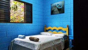 Een kleine, kleurrijke kamer met een enkel bed, een raam met uitzicht op begroeiing en een schilderij van een boot aan de muur.
