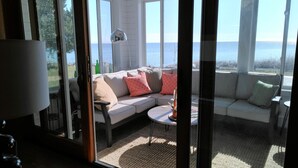 Ein sonnendurchflutetes Zimmer mit einem weißen Sofa, einem kleinen runden Tisch und Meerblick durch große Fenster.