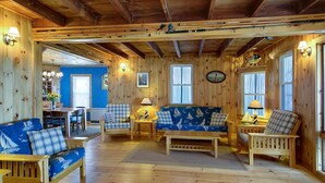 A cozy cabin interior with wooden walls, a blue sofa, a dining table, and a kitchen area visible through an open door. - The Boathouse Cottage is a beachfront family retreat (Saco)