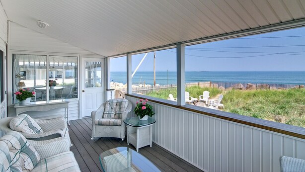 A covered outdoor seating area with wicker furniture, a glass table, and a view of the ocean. - The Boathouse Cottage is a beachfront family retreat (Saco)