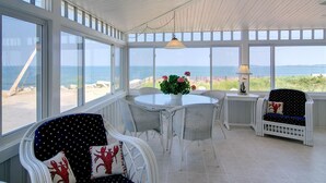 A sunlit room with large windows overlooking a beach, featuring wicker furniture and a table with a flower vase. - The Boathouse Cottage is a beachfront family retreat (Saco)