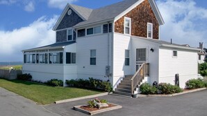 A two-story house with a large bay window, a wooden gable, and a front porch with steps. - The Boathouse Cottage is a beachfront family retreat (Saco)