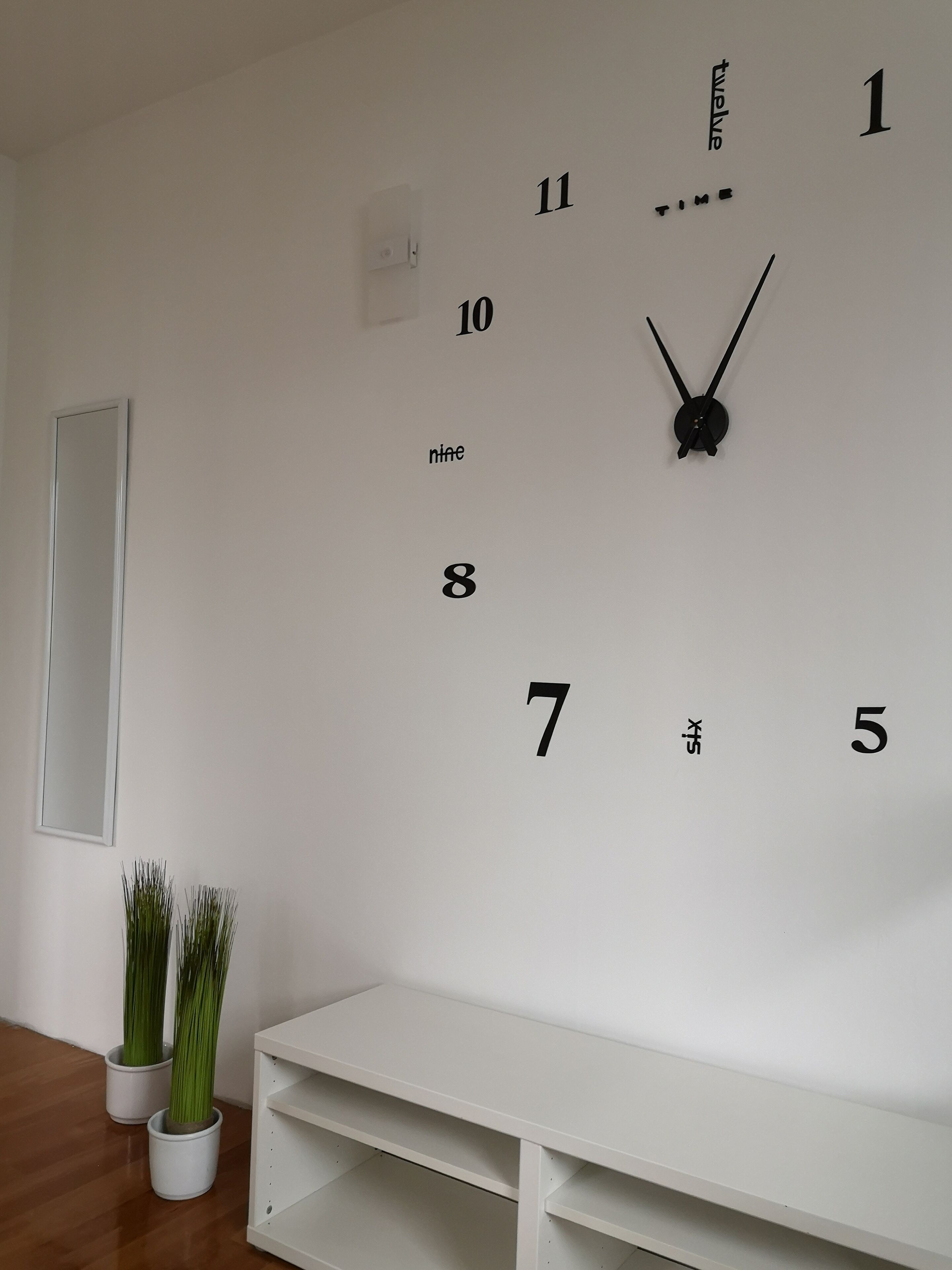 Eine schlichte Wanduhr mit Zahlen und Zeigern, ein weißes Regal und zwei Blumentöpfe.