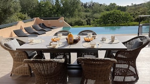 A dining table set for breakfast with wicker chairs, a view of a swimming pool, and lush greenery in the background.
