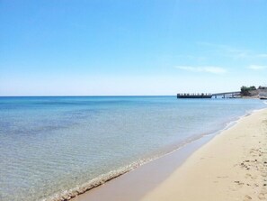 A beach with clear water, a sandy shore, and a pier in the distance.