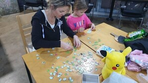 Due persone sedute a un tavolo di legno con tessere di un puzzle, un peluche di Pikachu e un portatile.