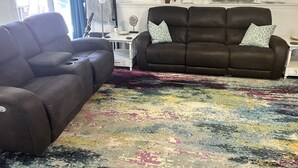 Un salon avec un canapé marron et un fauteuil marron, un tapis coloré, une table d’appoint avec une lampe et une fenêtre ornée de plantes en pot.