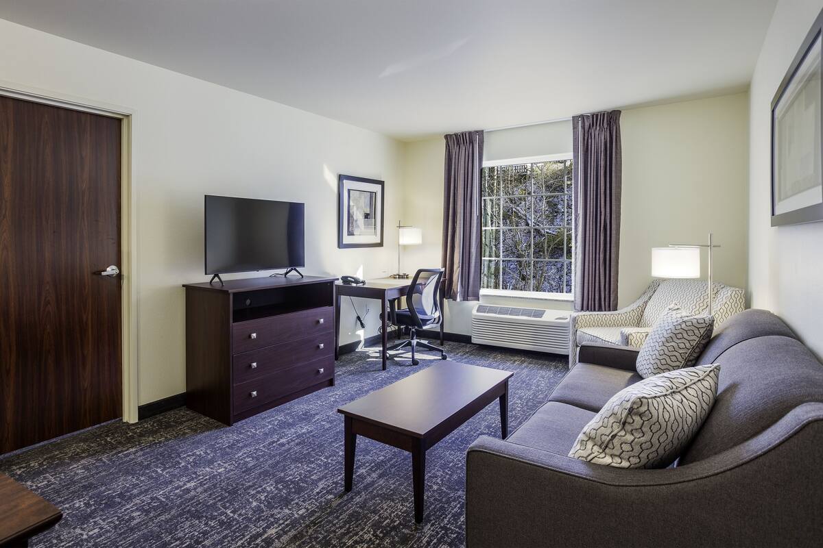 A hotel room with a dark wood wardrobe, a flat-screen TV, a desk with a chair, a sofa with patterned cushions, and a large window with curtains.