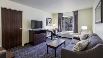 Ein Hotelzimmer mit dunklen Holzschränken, einem Flachbildfernseher, einem Schreibtisch mit Stuhl, einer Couch mit gemusterten Kissen und einem großen Fenster mit Vorhängen.