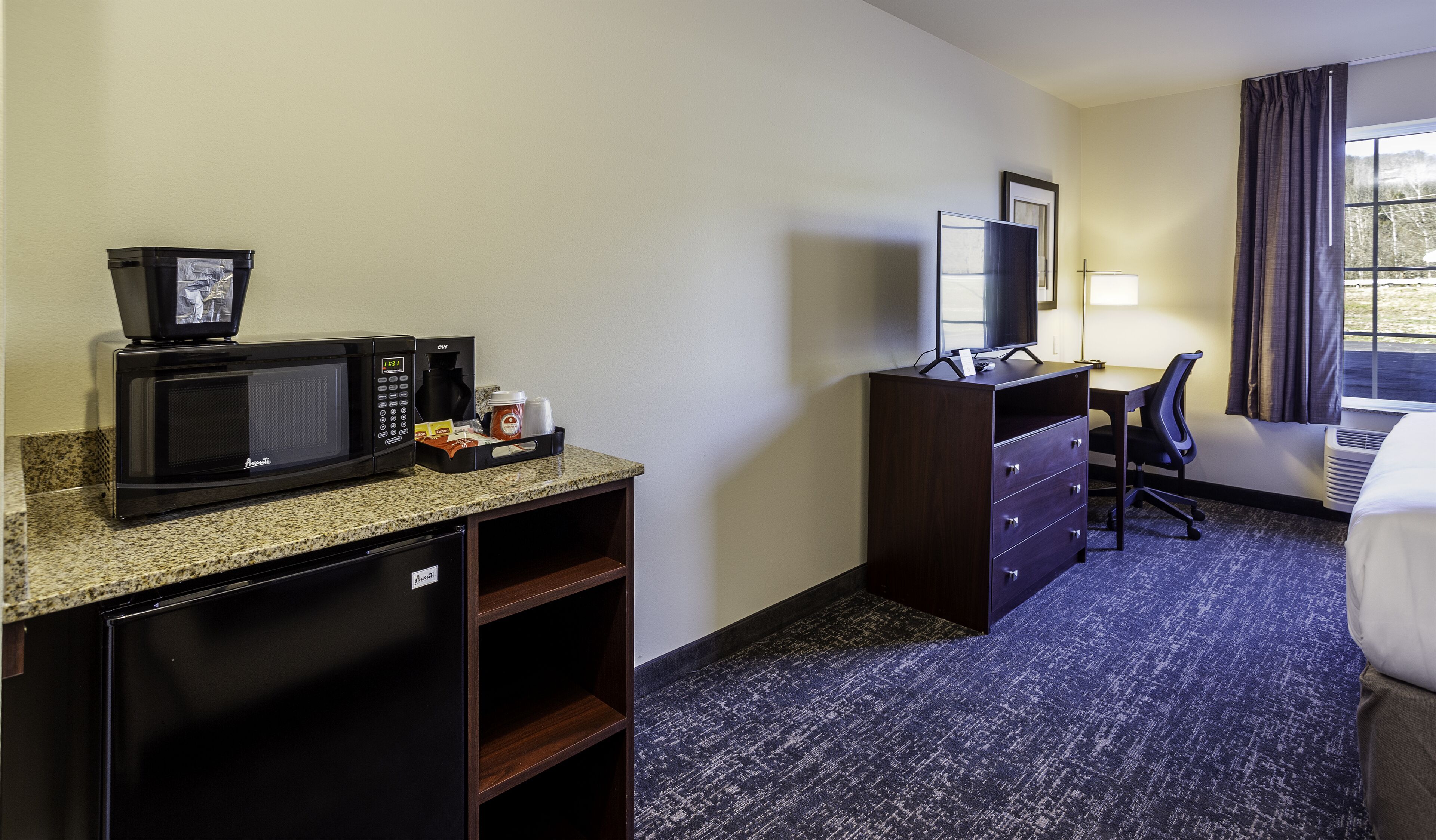 Une chambre d’hôtel équipée d’une kitchenette, d’un bureau, d’une chaise, d’un lit et d’une fenêtre avec des rideaux.
