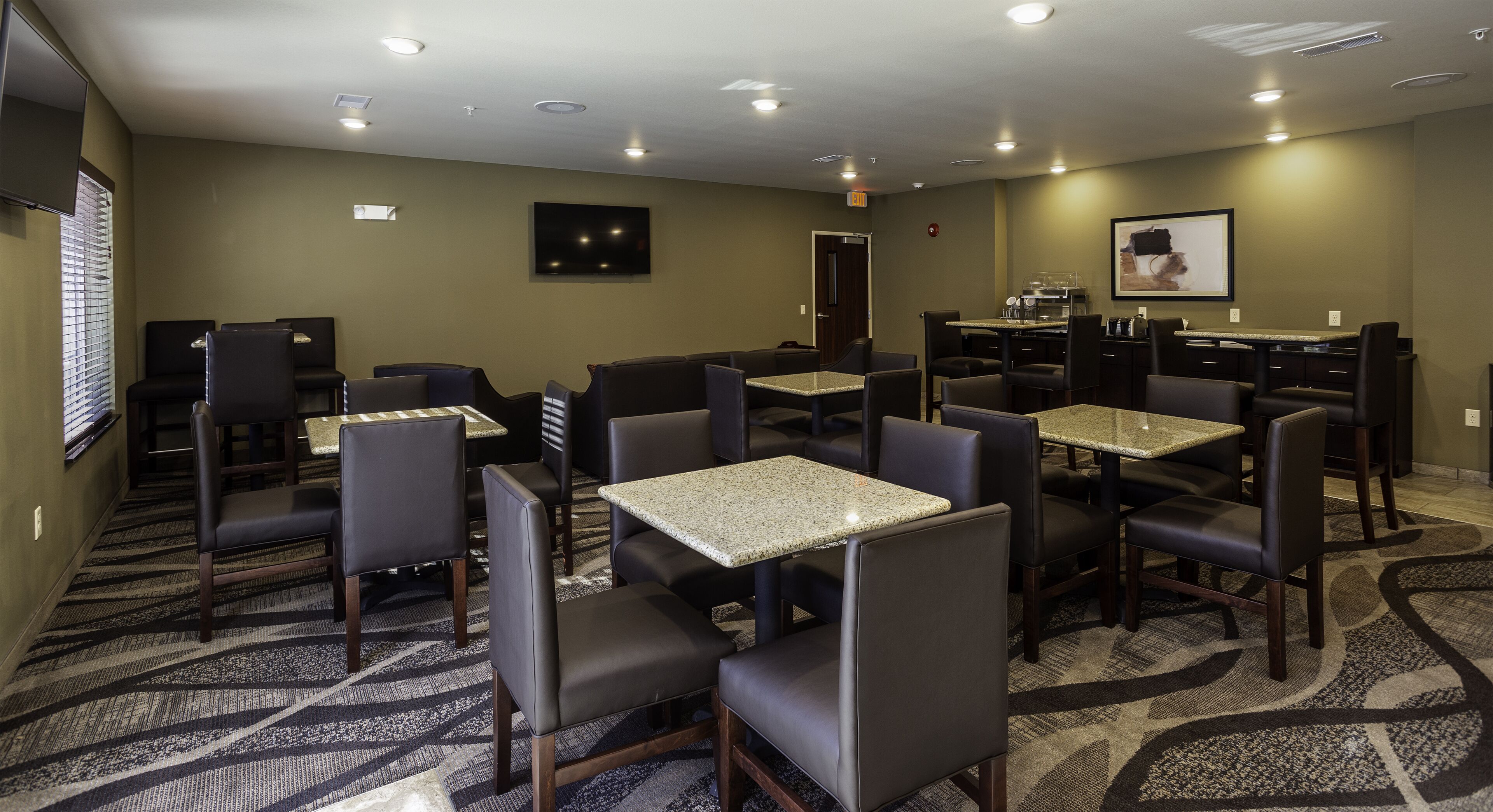 A hotel dining area with multiple tables and chairs, a television mounted on the wall, and a patterned carpet.