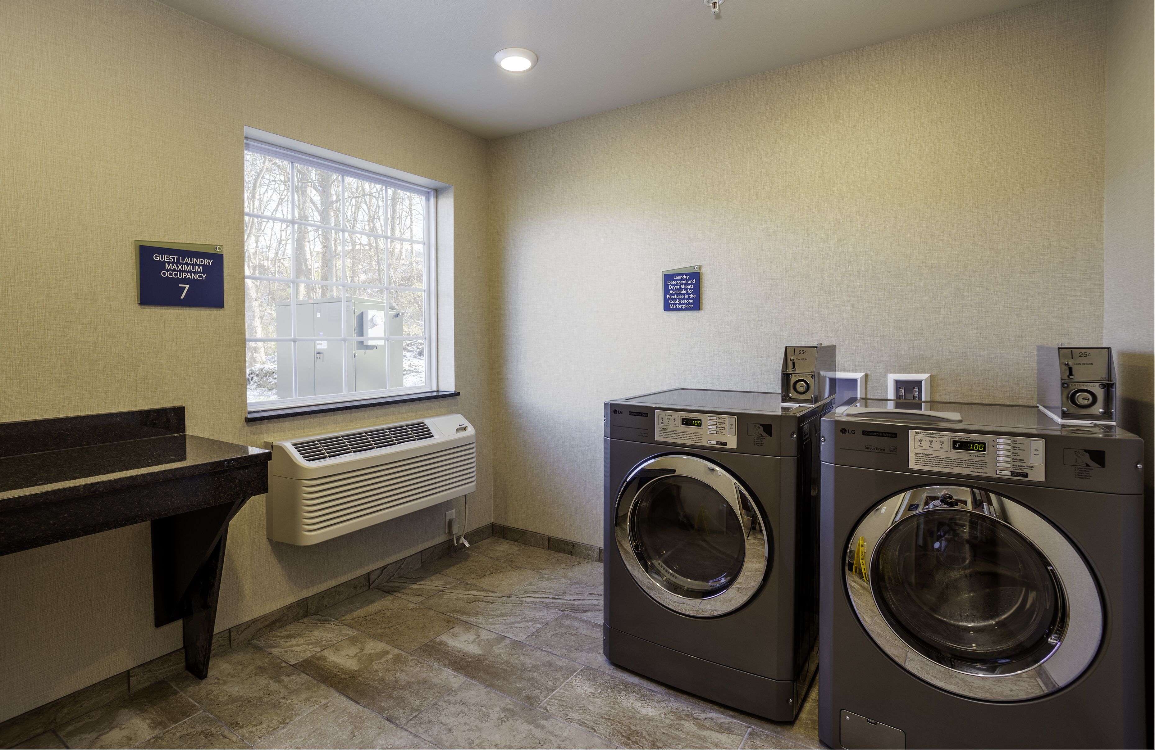 A laundry room with two front-loading washing machines, a bench, a window, and a sign indicating guest laundry maximum occupancy.