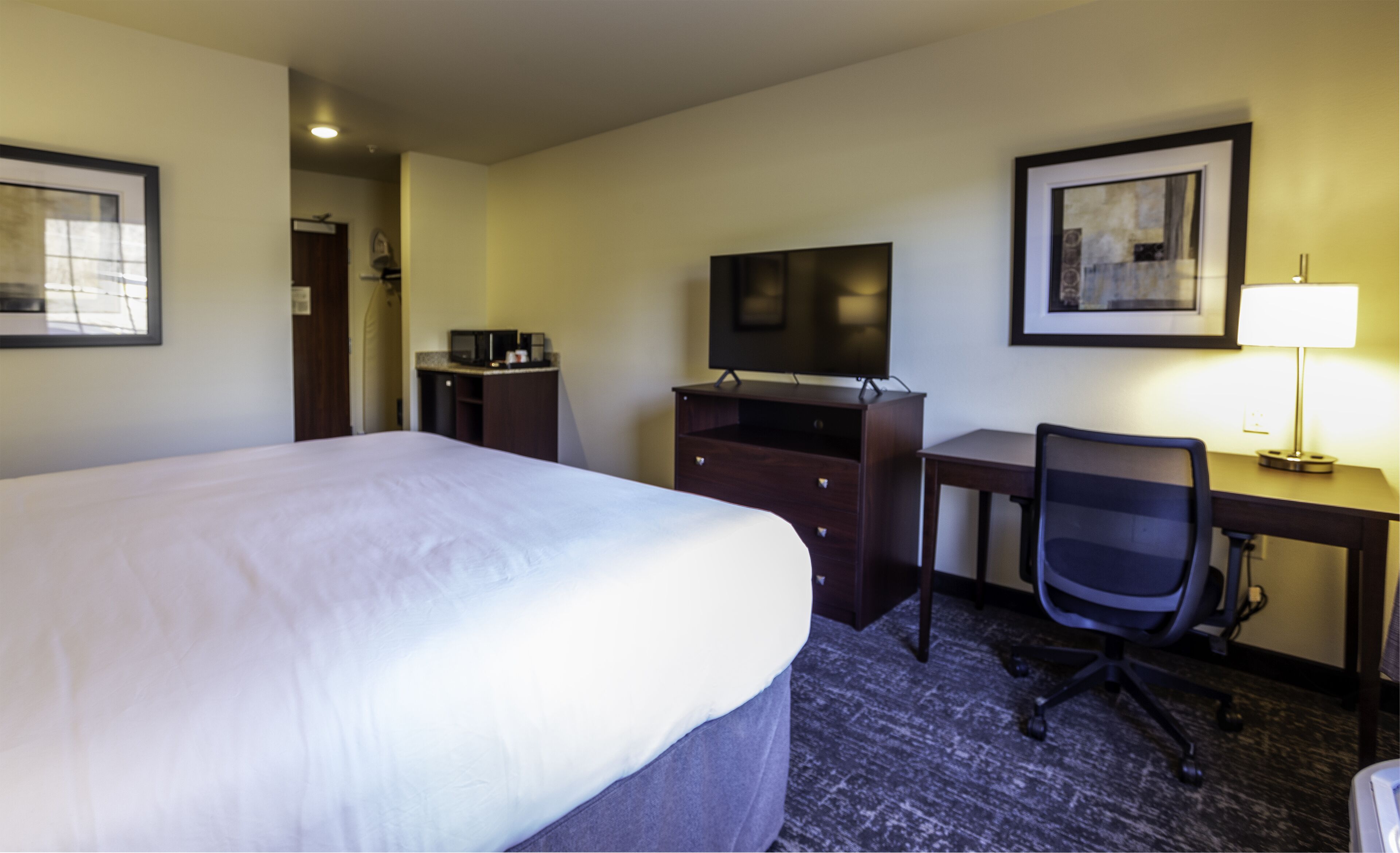 Ein Hotelzimmer mit einem großen Bett, einem Schreibtisch mit Computer, einem Sessel, einem Fernseher und einer Lampe.