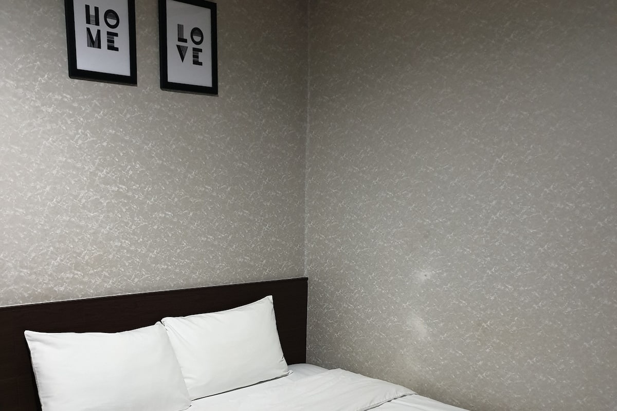 A neatly made bed with white linens in a room with textured gray walls. Two framed signs on the wall read "HOME" and "LOVE".