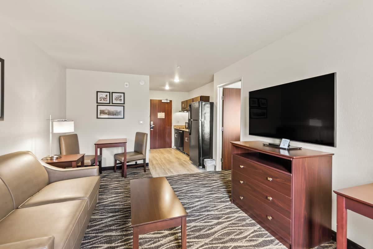 A modern hotel room with a sofa, a TV, a dining area, and a kitchen.