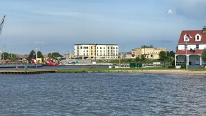 A waterfront view with a dock, a building with multiple windows, and a flag on a house.