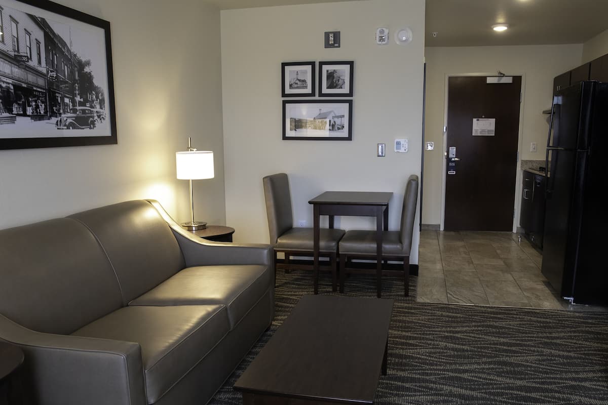 A modern hotel room with a sofa, a small table, a dining area with chairs, a kitchen with a refrigerator, and framed pictures on the wall.