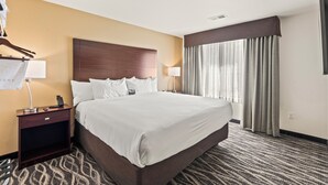 A hotel room with a large bed, a wooden headboard, two bedside lamps, a nightstand, a hanging coat rack, and a window with curtains. - Cobblestone Hotel & Suites - Two Rivers (Two Rivers)