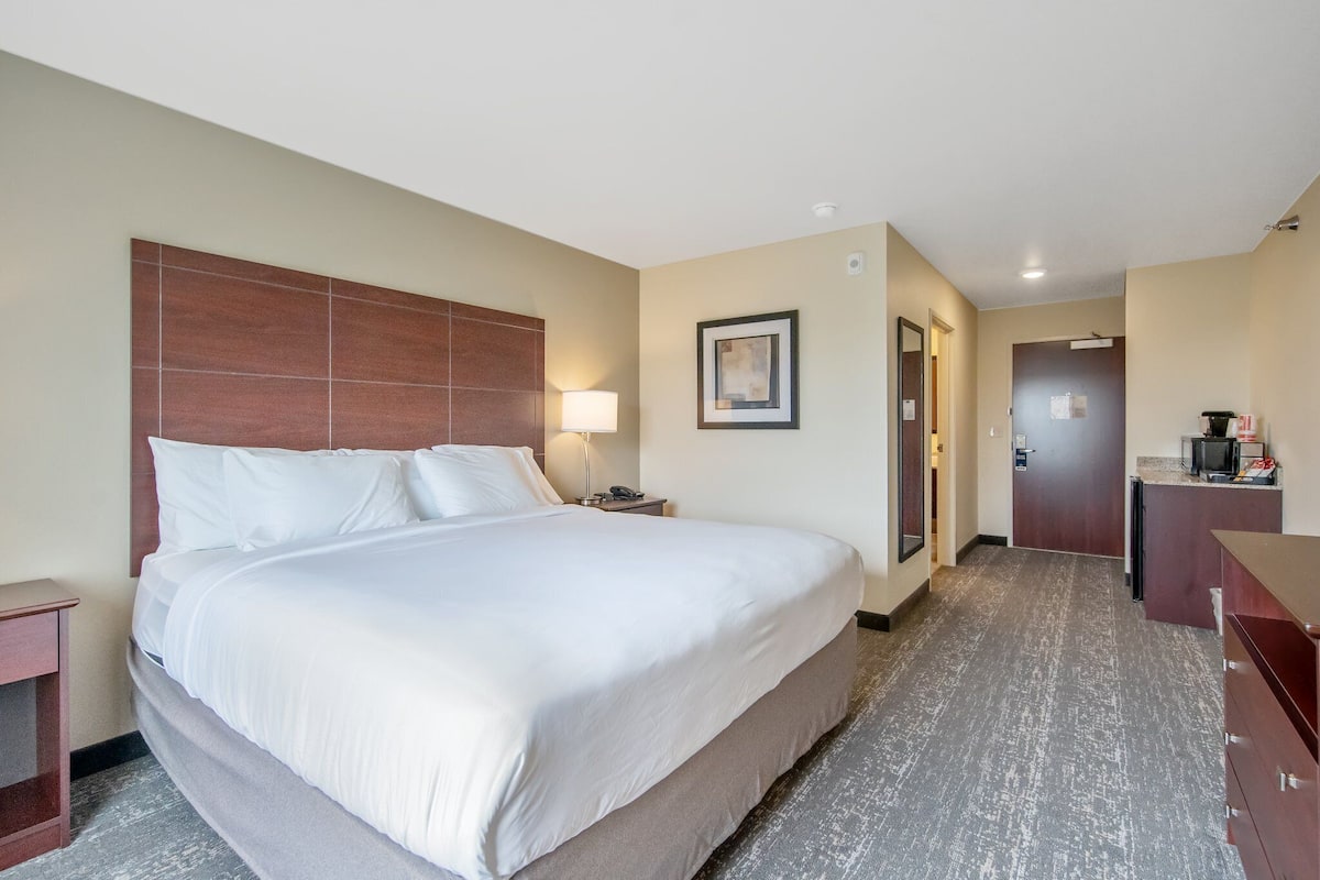 A hotel room with a large bed, a nightstand, a desk with a lamp, and a door leading to a kitchenette.