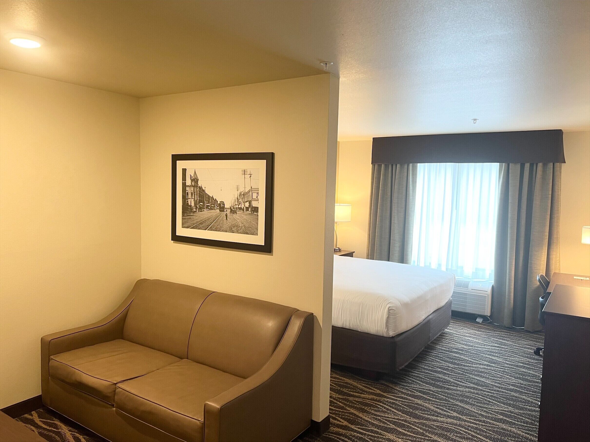 Habitación de hotel con sofá, cama, un cuadro enmarcado en la pared y una ventana con cortinas.