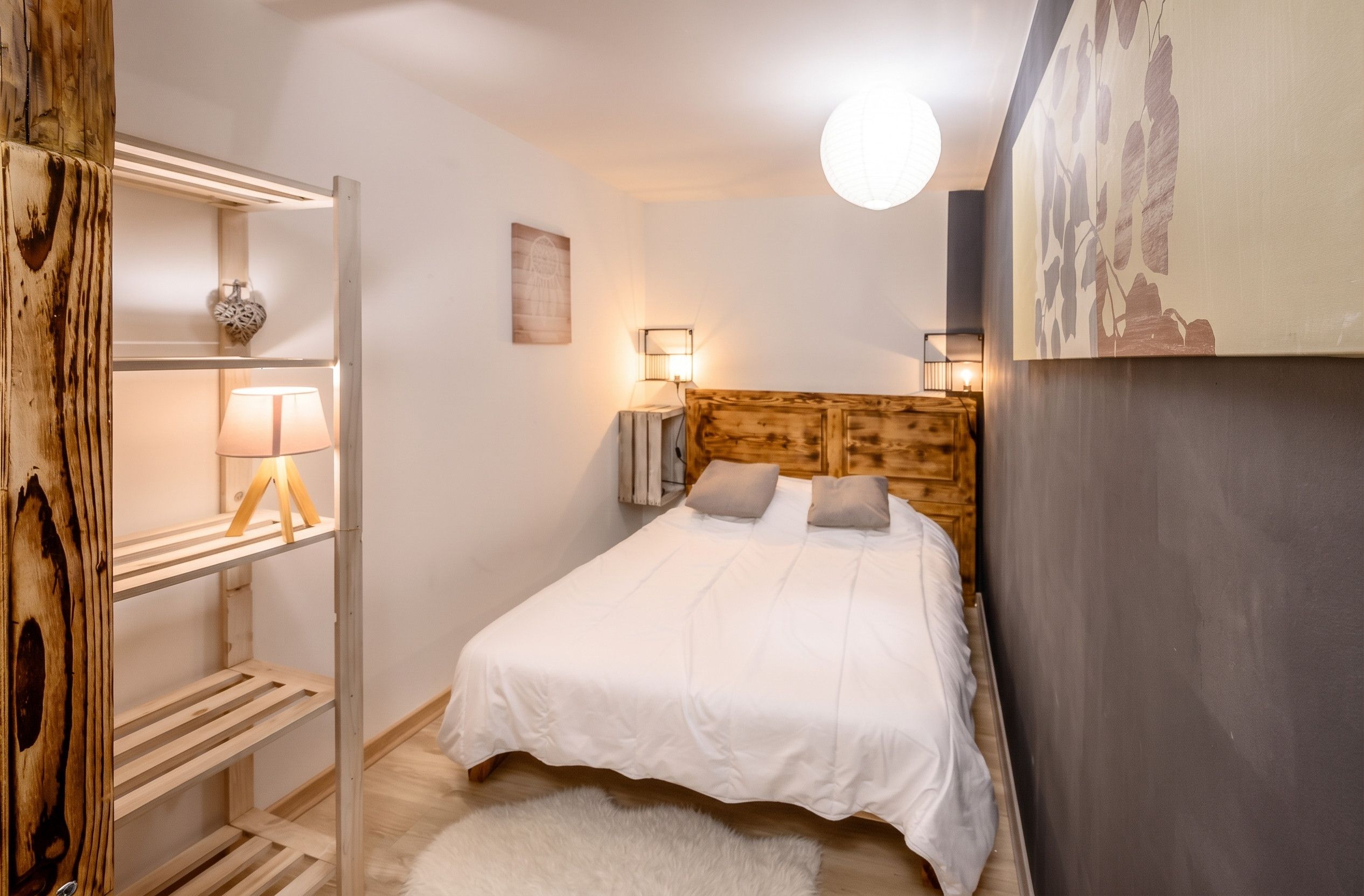 A bedroom with a wooden headboard, a white bed, two bedside lamps, and a wall-mounted shelf with a lamp.
