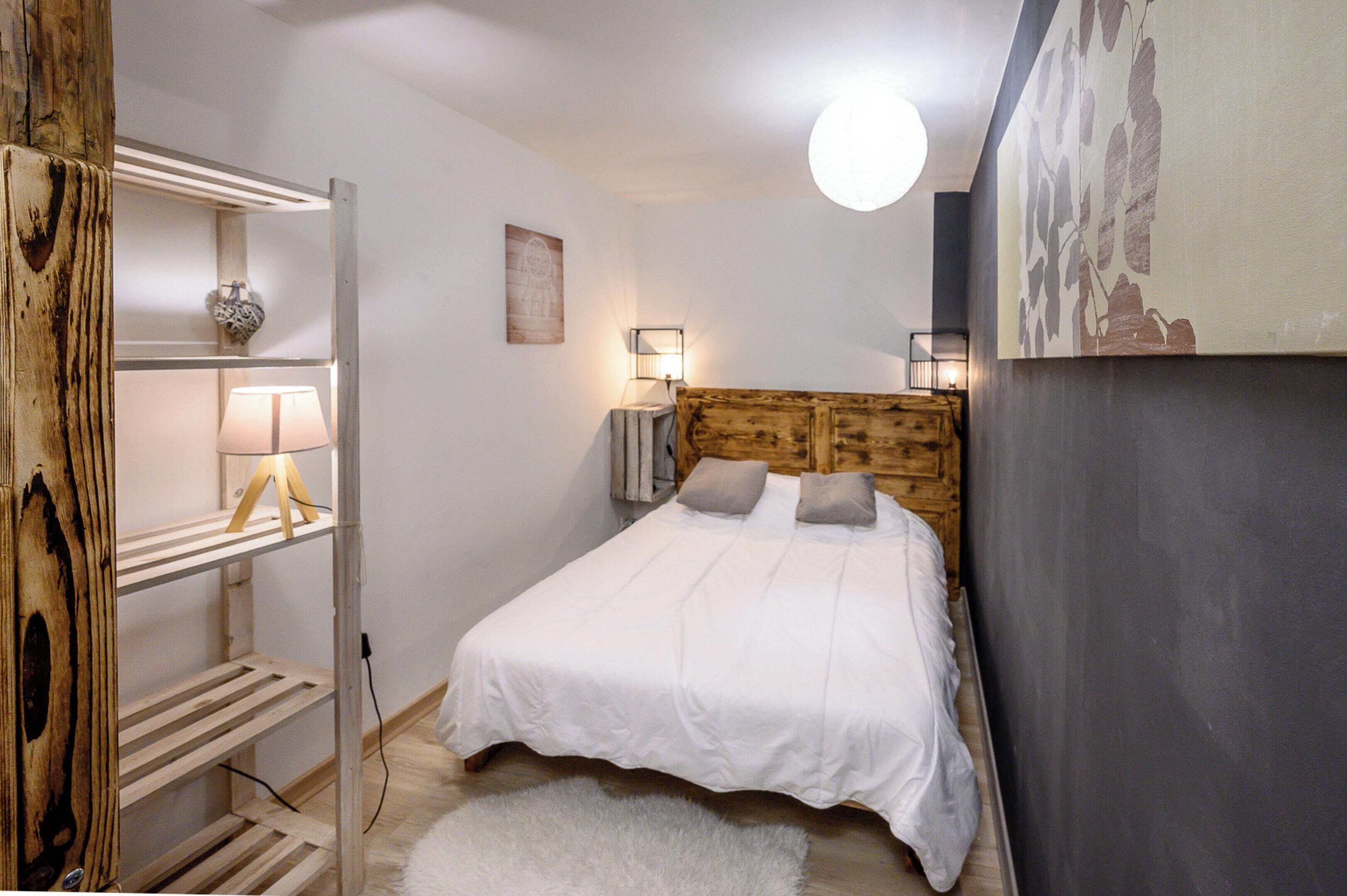 Un dormitorio con cabecera de madera, cubrecama blanco, dos almohadas, alfombra mullida blanca, estante con lámpara y una pintura mural.