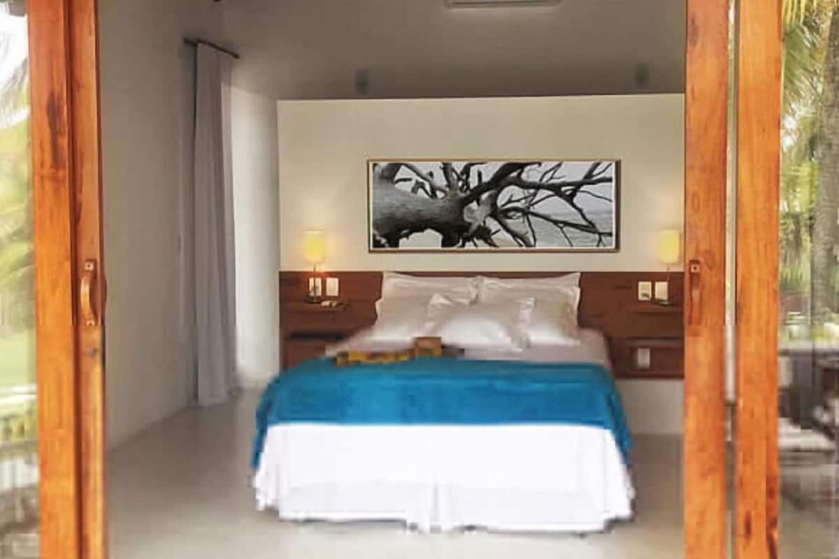 Ein Schlafzimmer mit einem Bett, weißer Bettwäsche, einer blauen Tagesdecke, einem Nachttisch mit Lampe und einem an der Wand hängenden Baumgemälde.