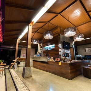 Un vestíbulo de hotel con un mostrador de recepción de madera, grandes lámparas colgantes y un reloj en la pared.