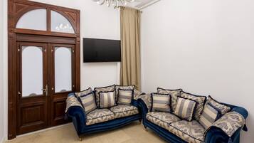 A living room with a dark blue sofa set, a wooden door with glass panels, a flat-screen TV mounted on the wall, and a chandelier.