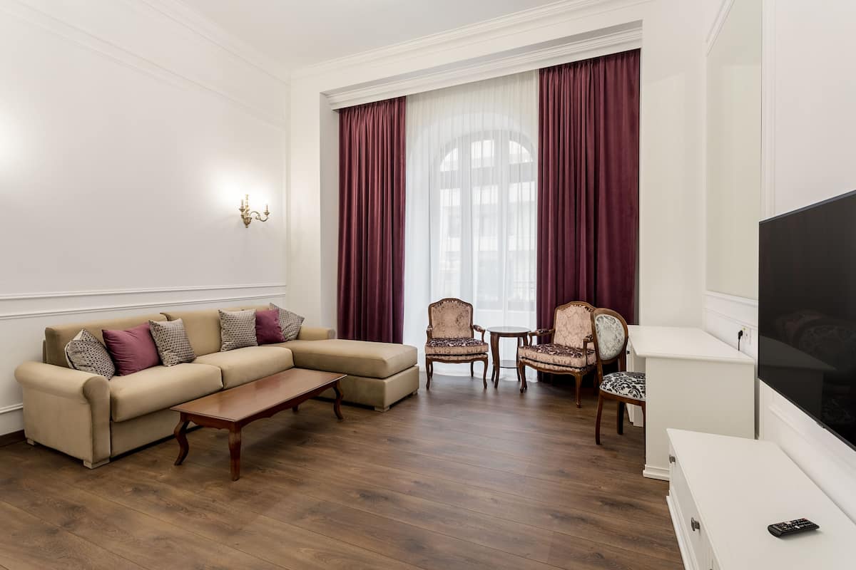 Ein Wohnzimmer mit einem beigefarbenen Sofa, gemusterten Sesseln, einem Holztisch und einem Fernseher auf einem weißen Schrank.