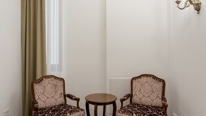 Une pièce comprenant deux fauteuils richement décorés, une petite table ronde, un lustre et une fenêtre avec des rideaux.