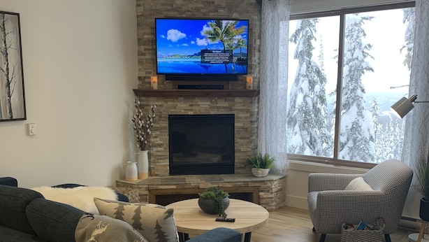 A living room with a stone fireplace, a flat-screen TV, a sofa, a chair, and a large window with a view of trees. - Updated house on the Ridge -3 suites, 1 hot tub open for summer, amazing views (Silver Star Mountain)