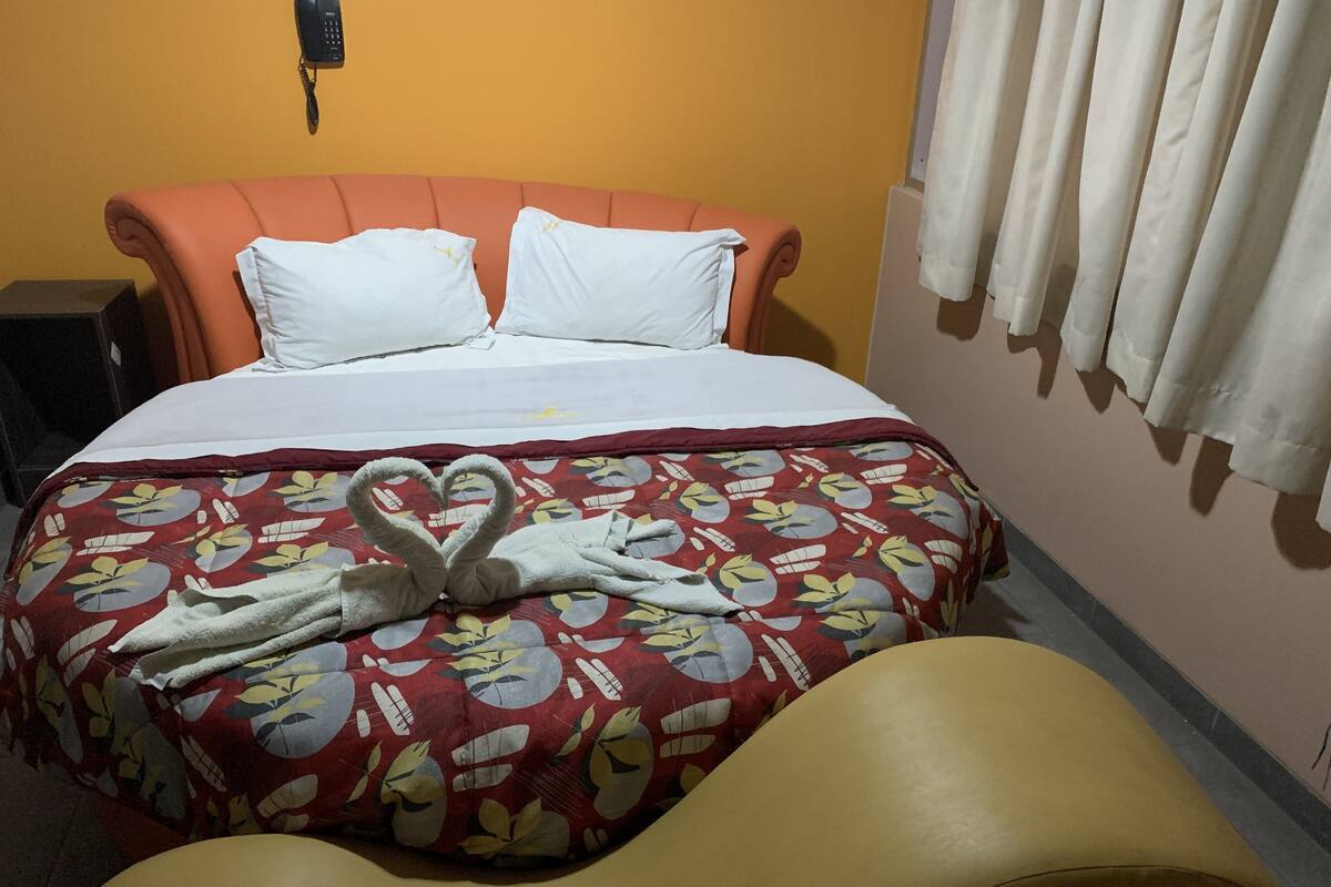 A hotel room with a bed, a red floral bedspread, a heart-shaped towel arrangement, and a yellow armchair.