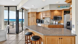 A modern kitchen with wooden cabinets, granite countertops, and stainless steel appliances. It features a large island with bar stools, a view of the ocean through large sliding glass doors, and a sign reading "Bench Blessings."