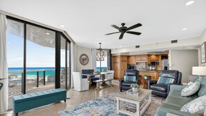 A spacious living room with a large window overlooking the ocean, a dining area with a table and chairs, a kitchen with wooden cabinets, and a seating area with a sofa and armchairs.