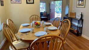 Uma sala de jantar com mesa de madeira posta para seis pessoas, rodeada por cadeiras de madeira. Há uma cesta de frutas sobre a mesa e um ventilador de teto.