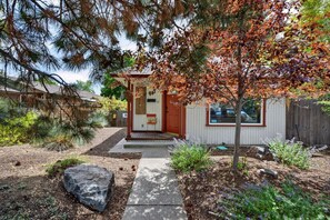 Une petite maison avec un porche, une terrasse en bois et un jardin agrémenté de plantes et d’un rocher.