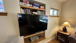 Uma sala de estar com uma TV de tela plana fixada em uma prateleira de madeira, uma mesinha com um abajur e uma janela com vista para árvores.
