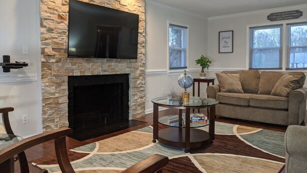 A living room with a stone fireplace, a flat-screen TV mounted above it, a glass coffee table, a brown leather sofa, and a patterned rug. - Lakeview Five Bedrooms Cape Cod Near Quantico and Historic Fredericksburg (Stafford Courthouse)