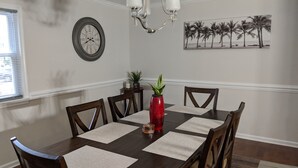 A dining room with a dark wooden table, four chairs, a red vase with a plant, a clock, and a beach-themed picture on the wall. - Lakeview Five Bedrooms Cape Cod Near Quantico and Historic Fredericksburg (Stafford Courthouse)
