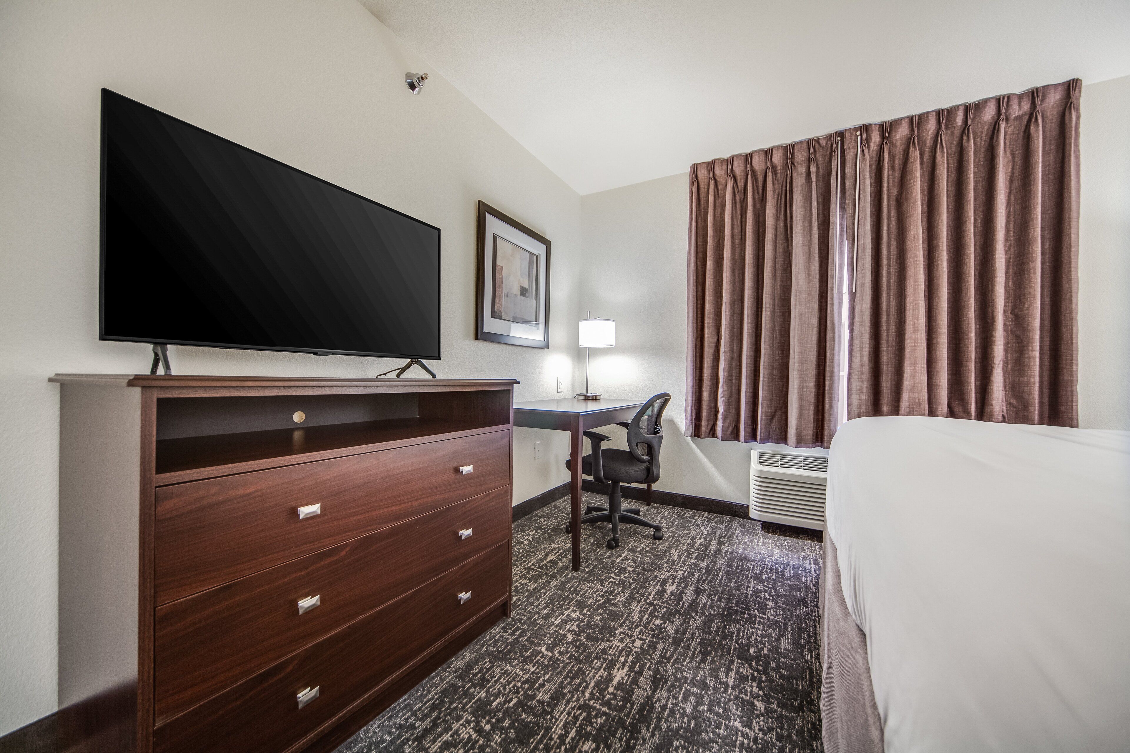 Hotelkamer met flatscreen televisie, bureau met stoel, bed, dressoir en raam met gordijnen.