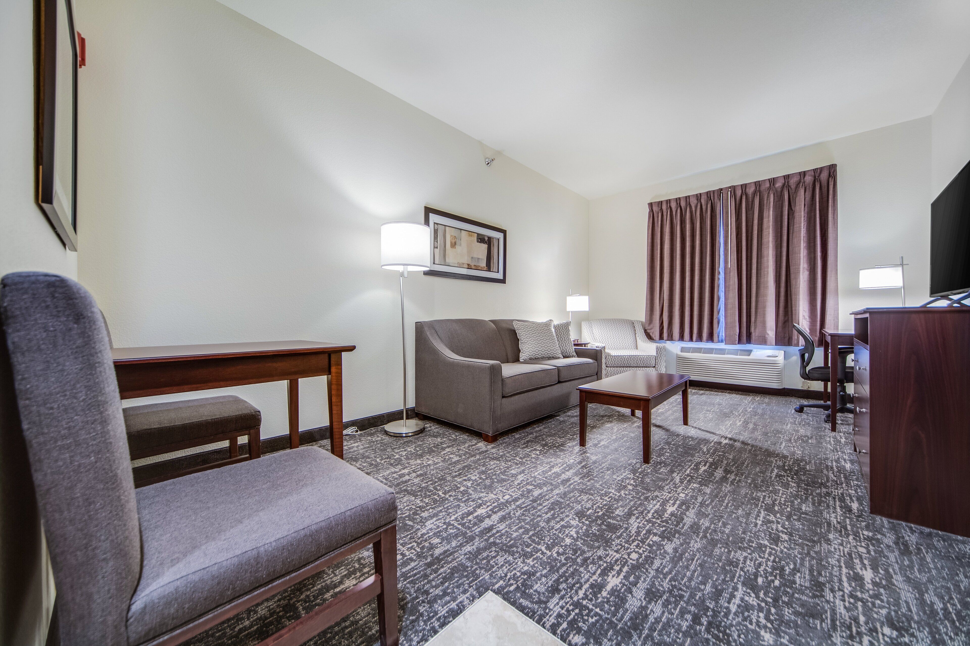 Une chambre d’hôtel avec un canapé gris, un bureau en bois, une petite table, un lampadaire, un téléviseur à écran plat et une grande fenêtre avec des rideaux.