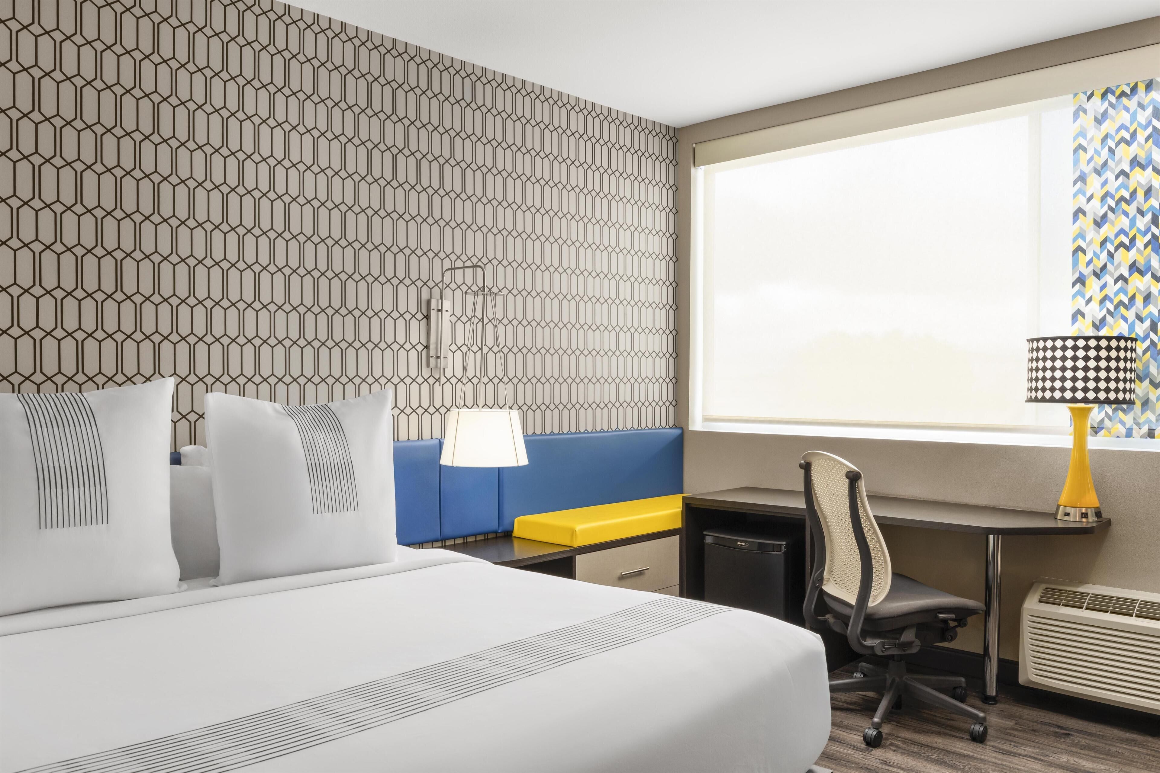 A hotel room with a large bed, a desk, a chair, a small table, a lamp, and a window with a view.