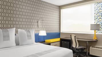 A hotel room with a large bed, a desk, a chair, a small table, a lamp, and a window with a view.