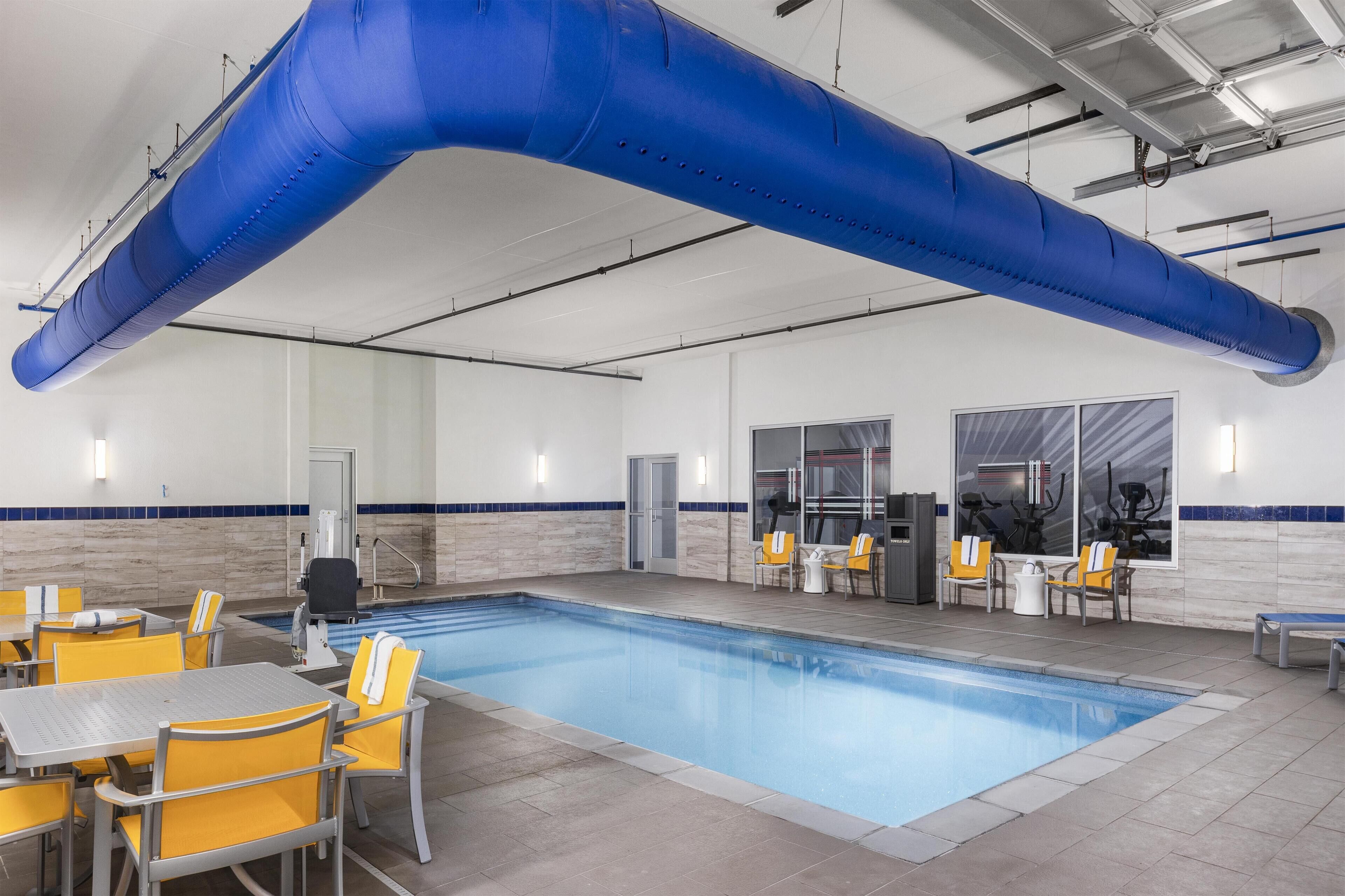 A swimming pool with yellow chairs and tables, a blue ceiling feature, and a door leading to another room.