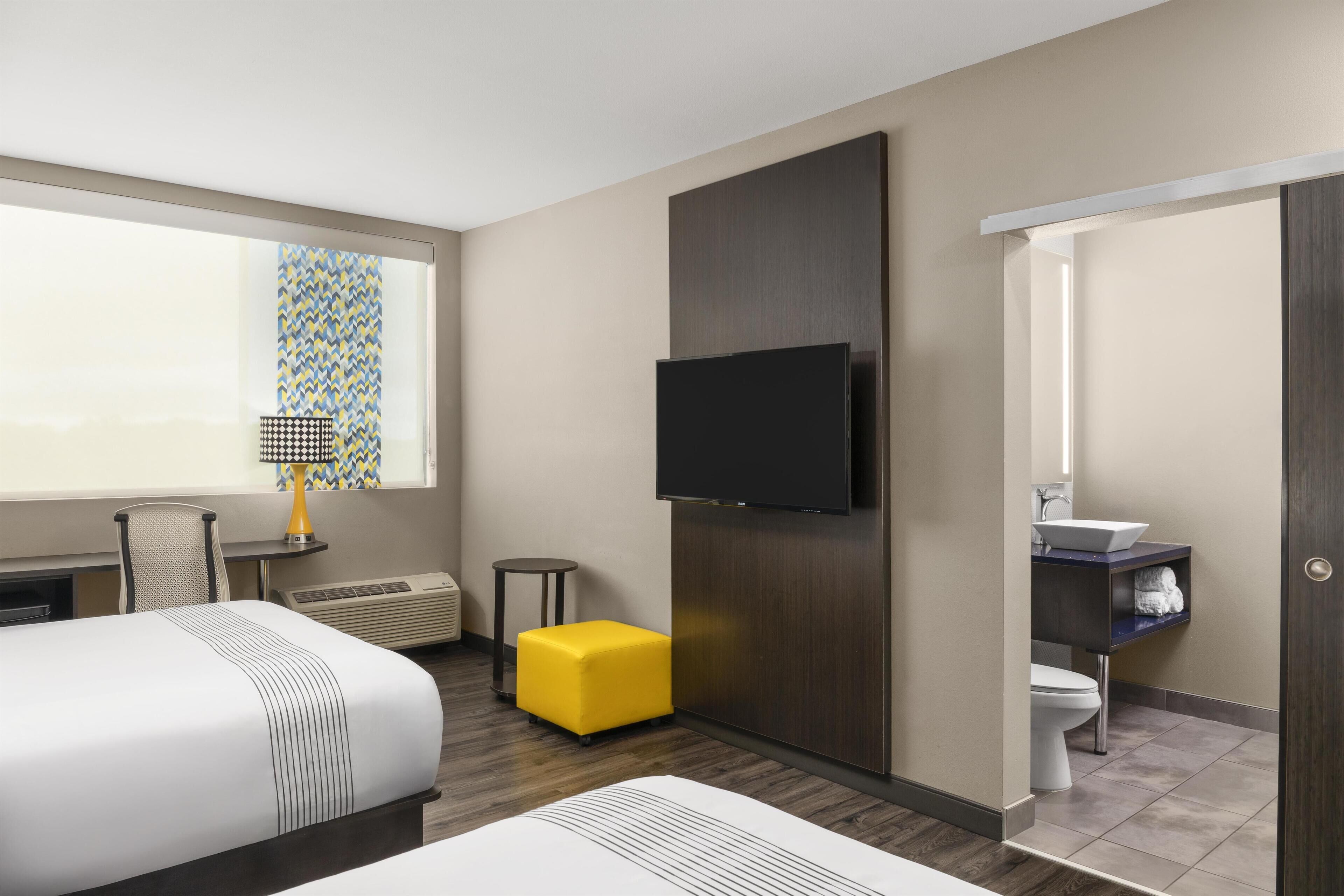 Habitación de hotel con cama, televisión montada en la pared, mesita de noche con lámpara, un reposapiés amarillo y baño con lavamanos e inodoro.
