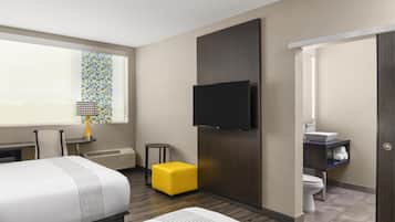 Une chambre d’hôtel avec un lit, un téléviseur fixé au mur, une petite table avec une lampe, un pouf jaune et une salle de bain avec un lavabo et des toilettes.