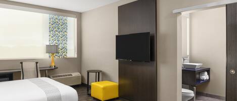 Habitación de hotel con cama, televisión montada en la pared, mesita de noche con lámpara, un reposapiés amarillo y baño con lavamanos e inodoro.